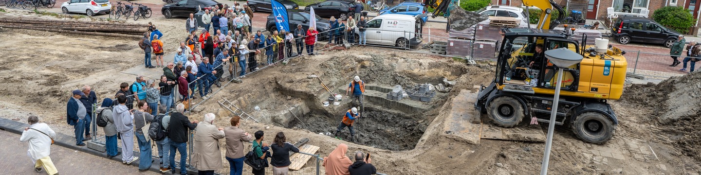 Geïnteresseerden staan rondom een opgravingsput te kijken naar de archeologen aan het werk.